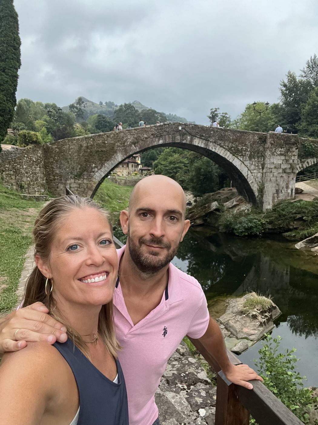 Melanie and her husband smiling in front of an old stone bridge over a green river